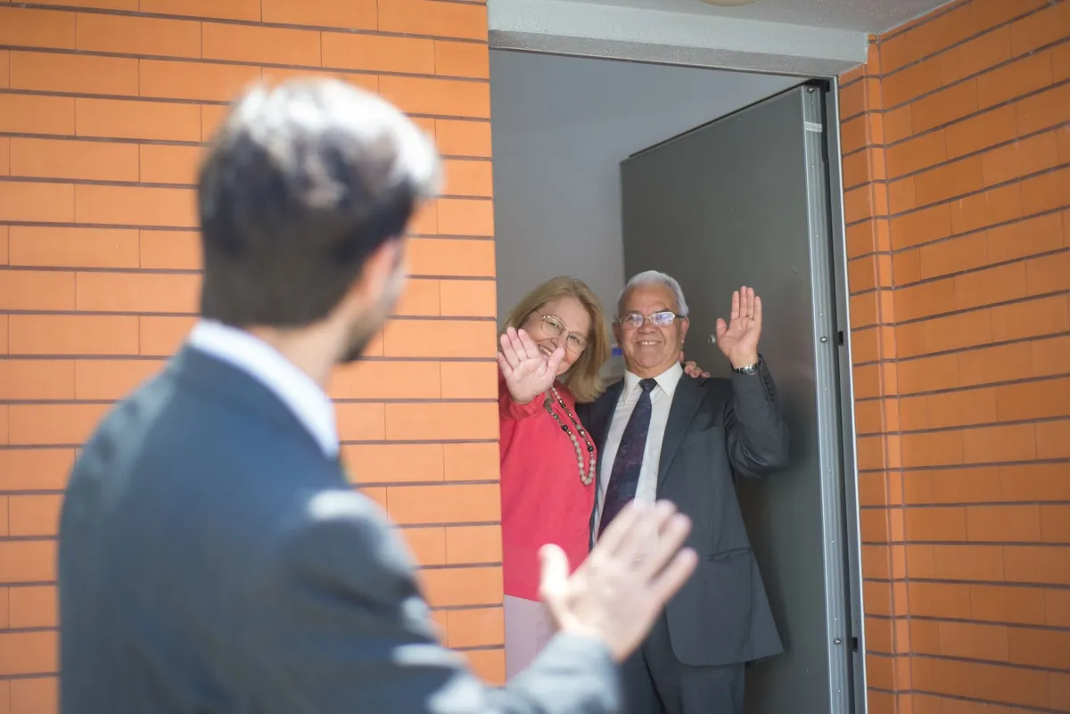 A man in a suit waving to an older couple standing in their front doorway