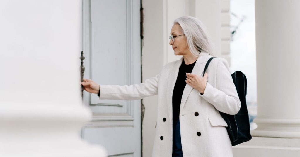 An older woman in a white coat opening the front door of a house