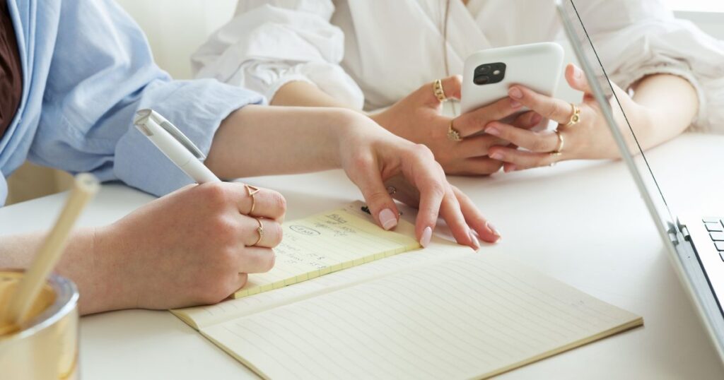 Two people at a white table, one writing in a notebook while the other holds a phone