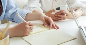 Two people at a white table, one writing in a notebook while the other holds a phone