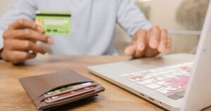 A person holding a bank card, back side up, next to a laptop and a leather card wallet