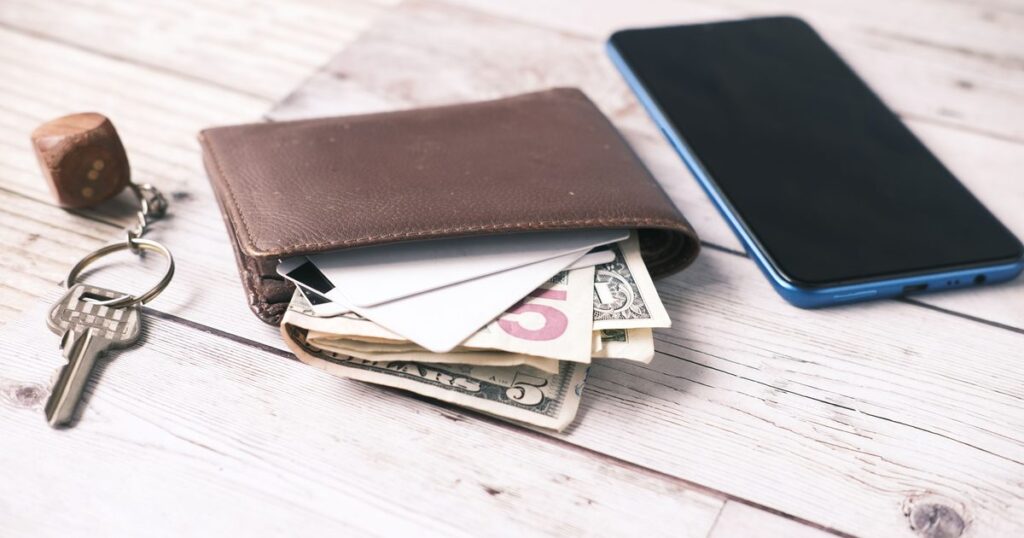A wallet with cash and cards beside a house key and a phone on a wooden table