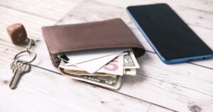 A wallet with cash and cards beside a house key and a phone on a wooden table