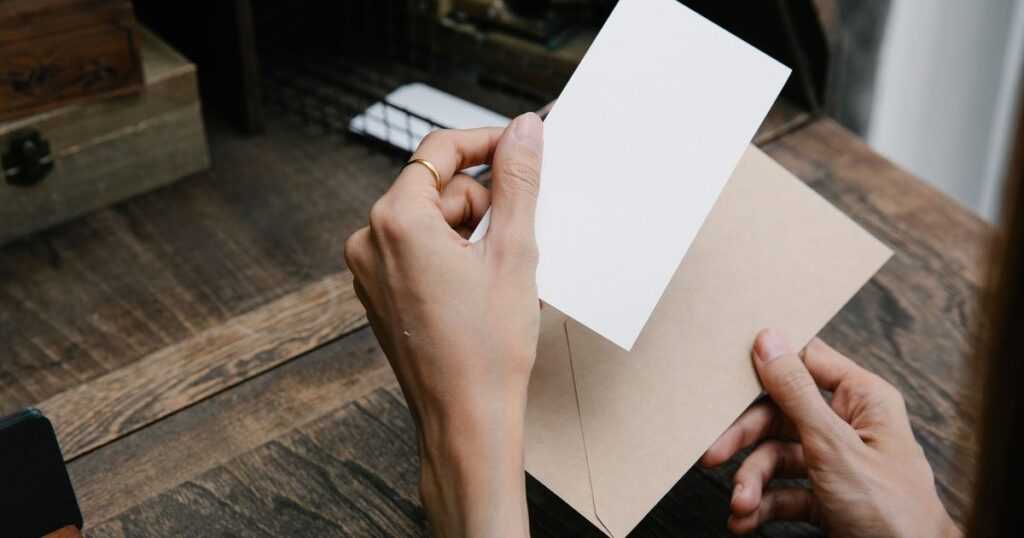 Two hands holding a blank white card over a brown paper envelope on a wooden desk