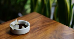 A white ceramic ashtray with ash on a wooden table, plants behind it