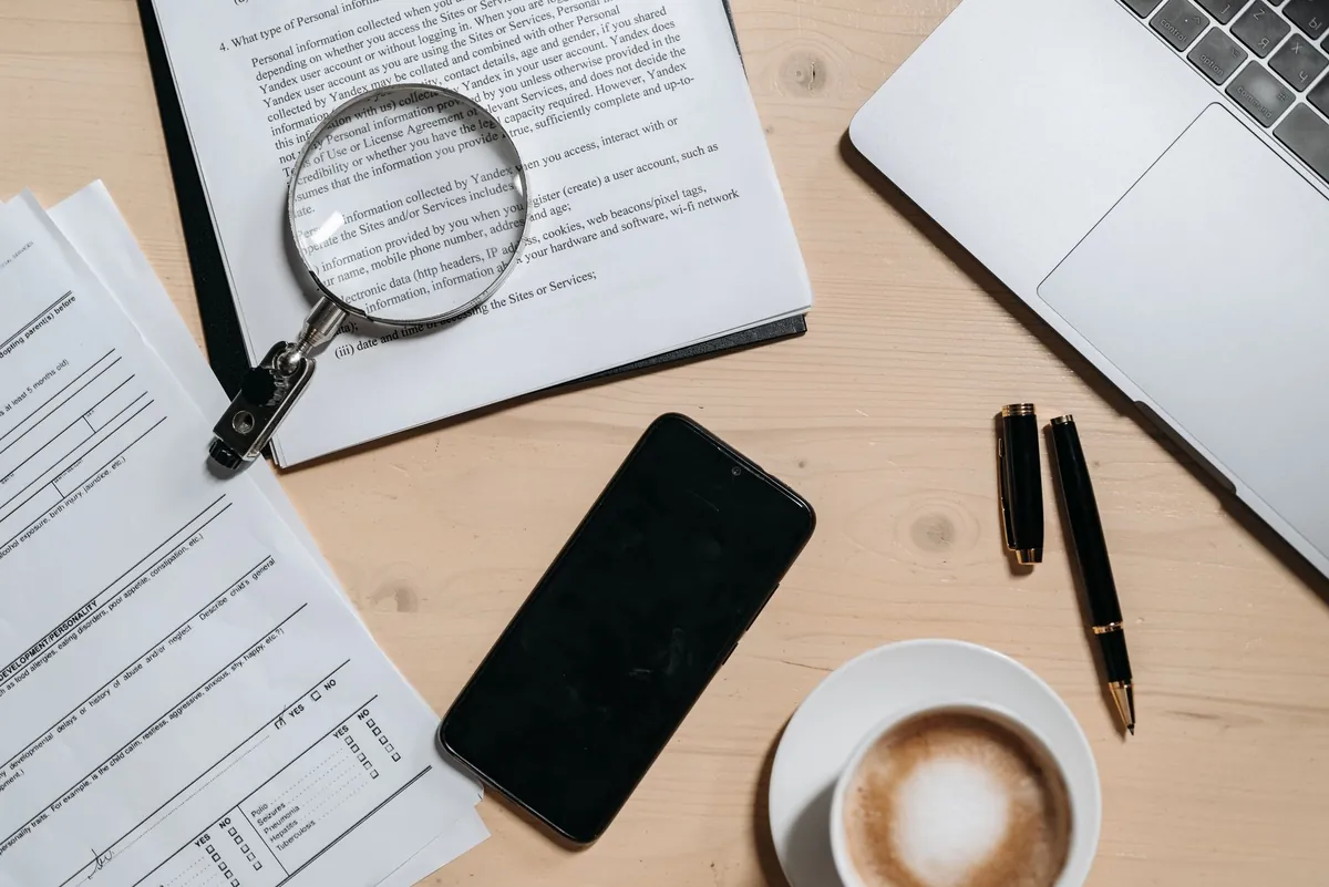 Printed documents, a magnifying glass, a phone and a cup of coffee laid out on a desk