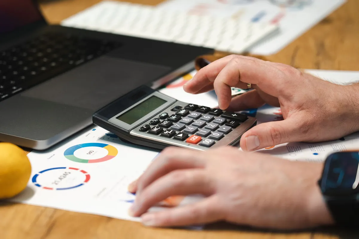 Hands working a desk calculator beside a printed chart and a laptop