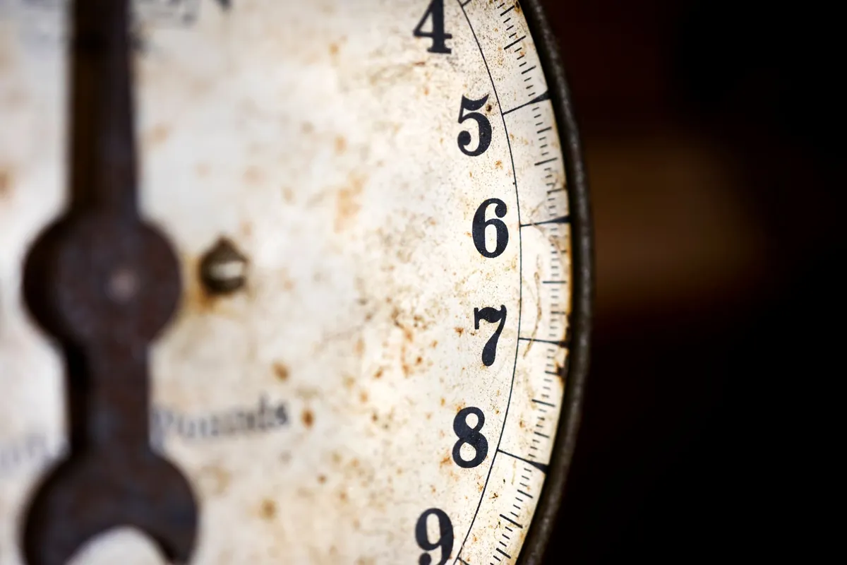 A macro of the rusted enamel face of an old weighing dial, the numbers 4 to 9 and graduation marks curving around a black pointer.