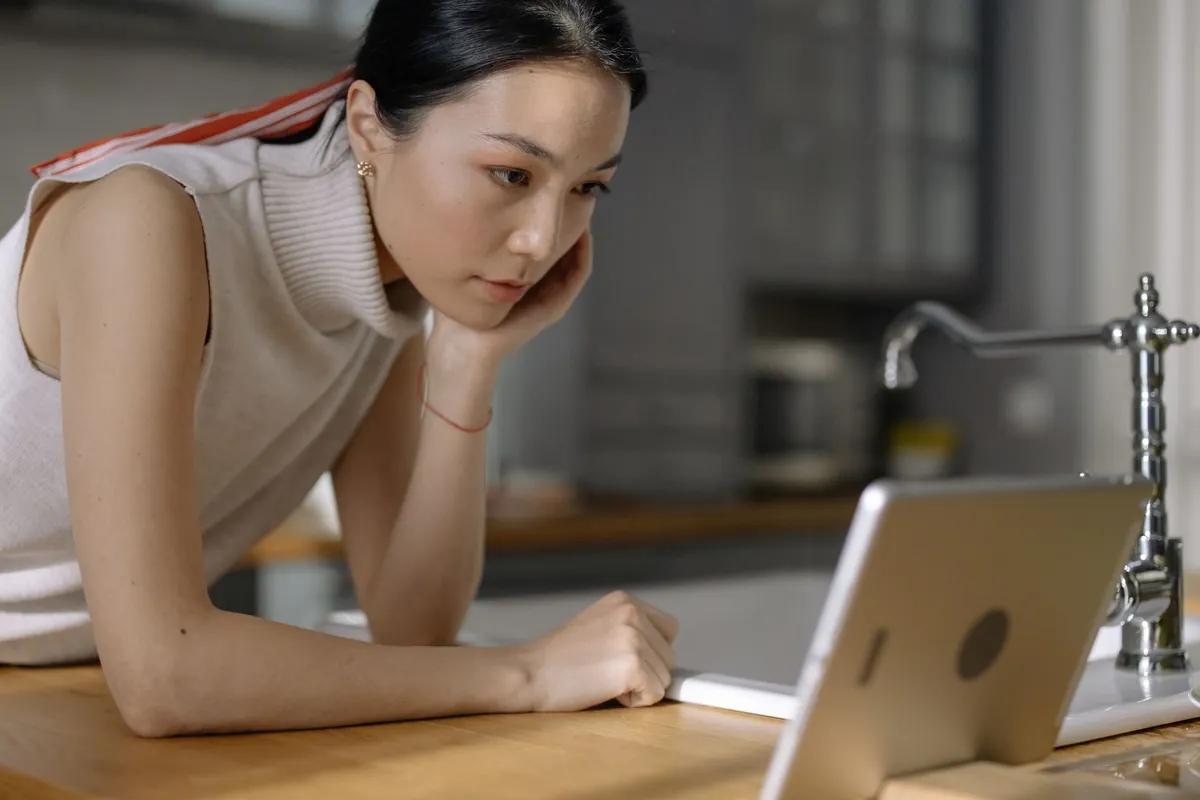 A woman leaning on a kitchen counter, reading closely from a tablet on a stand