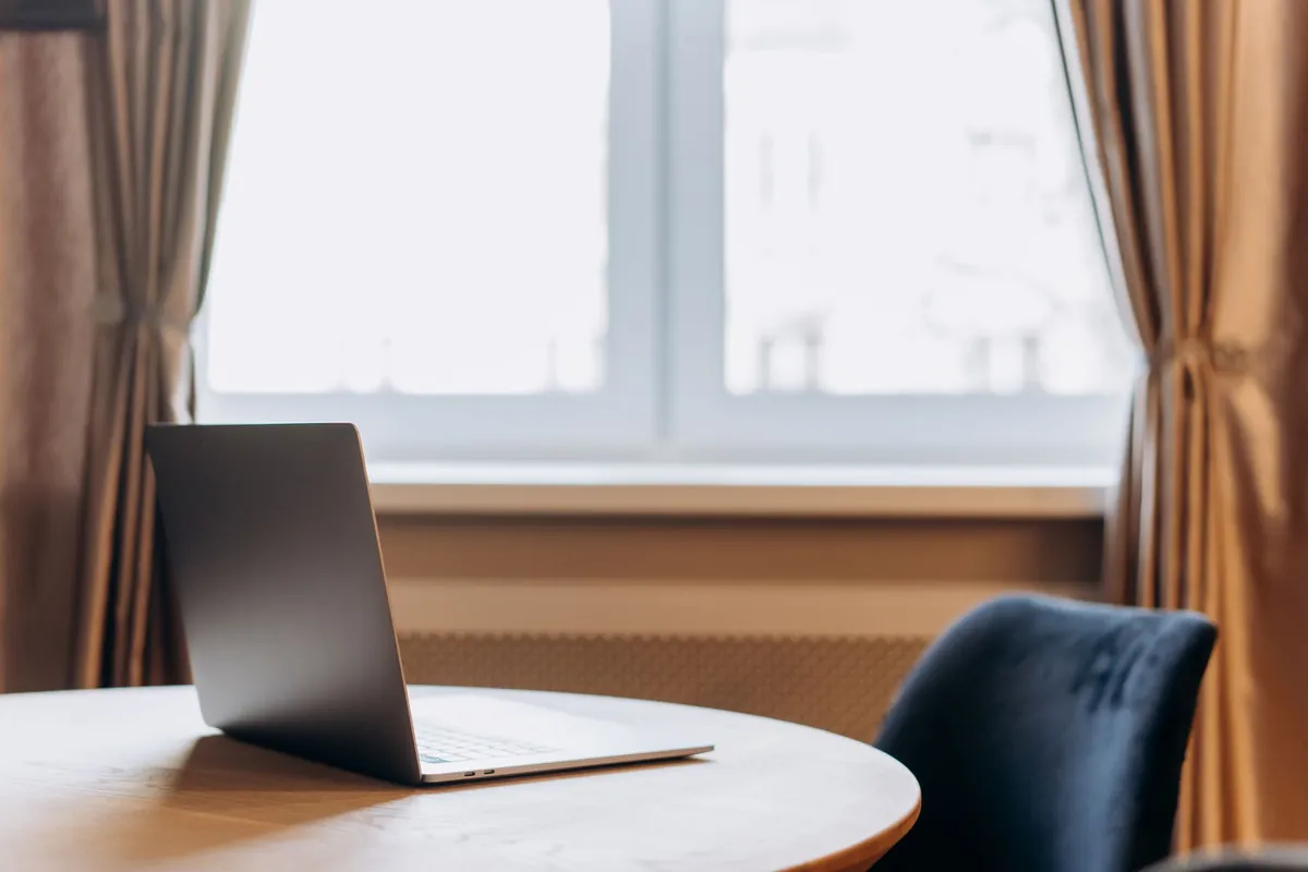 An open laptop on a round table beside a window, with an empty chair