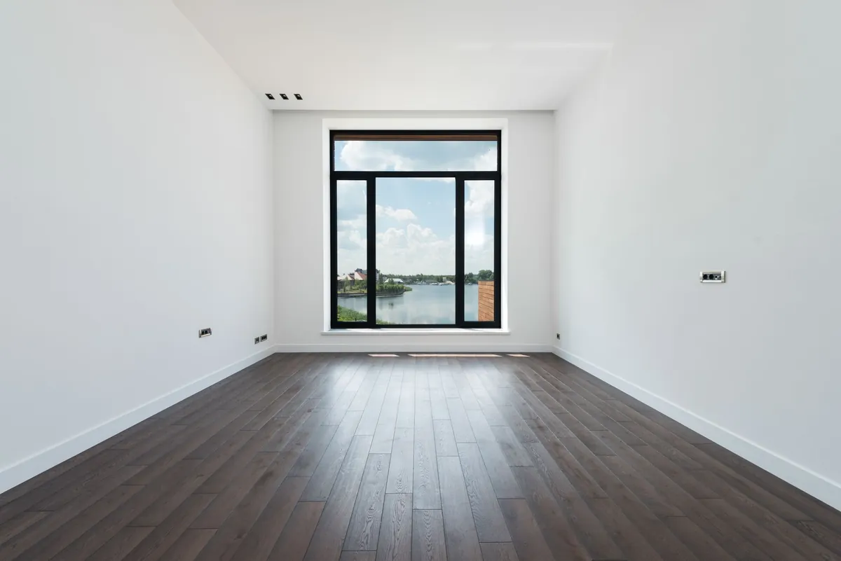 An empty room with wooden floors and a large window looking onto water