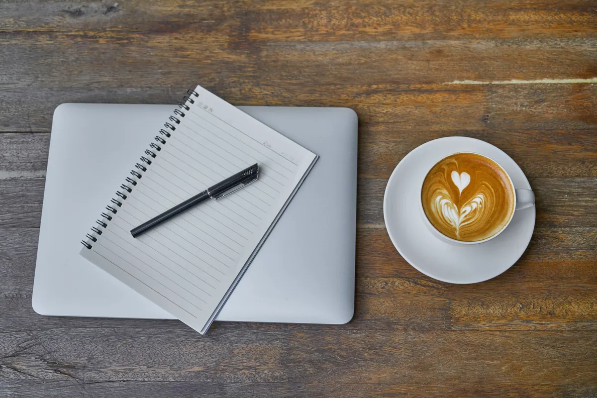 A coffee cup, a closed notebook, a pen and a laptop laid out on a rustic wooden table
