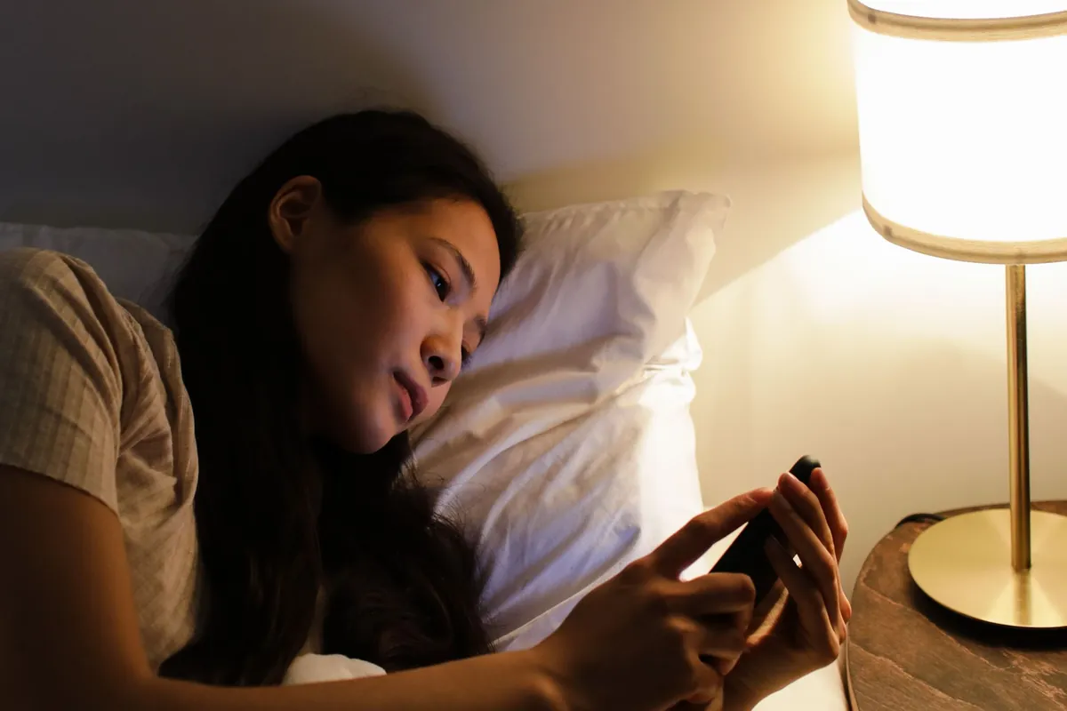 A woman lying in bed at night reading her phone by the light of a bedside lamp