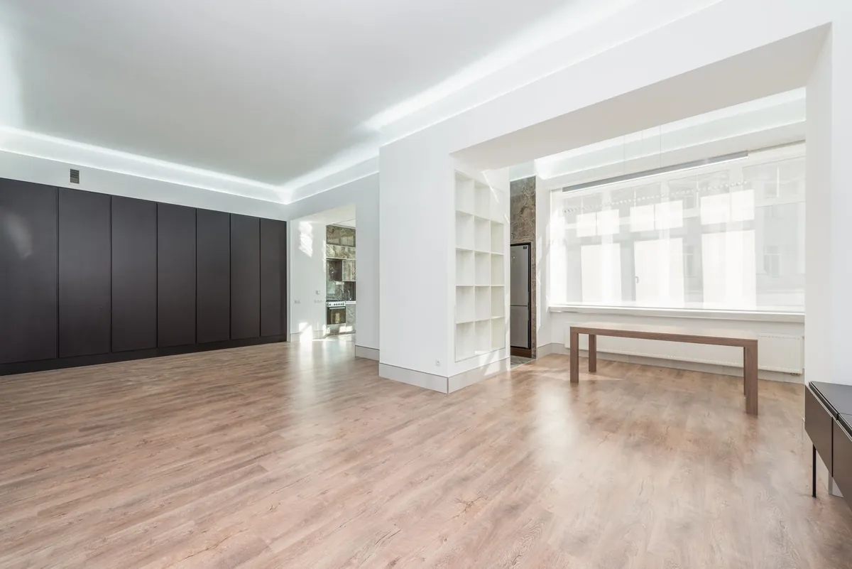 An empty apartment room with pale wooden floorboards, white walls and tall windows letting daylight fall across the floor.