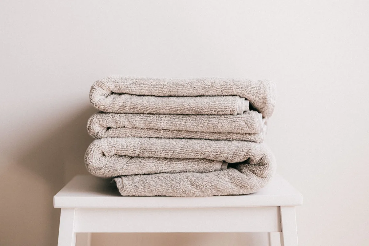 A stack of four folded beige bath towels sitting on a white wooden stool against a plain pale wall.