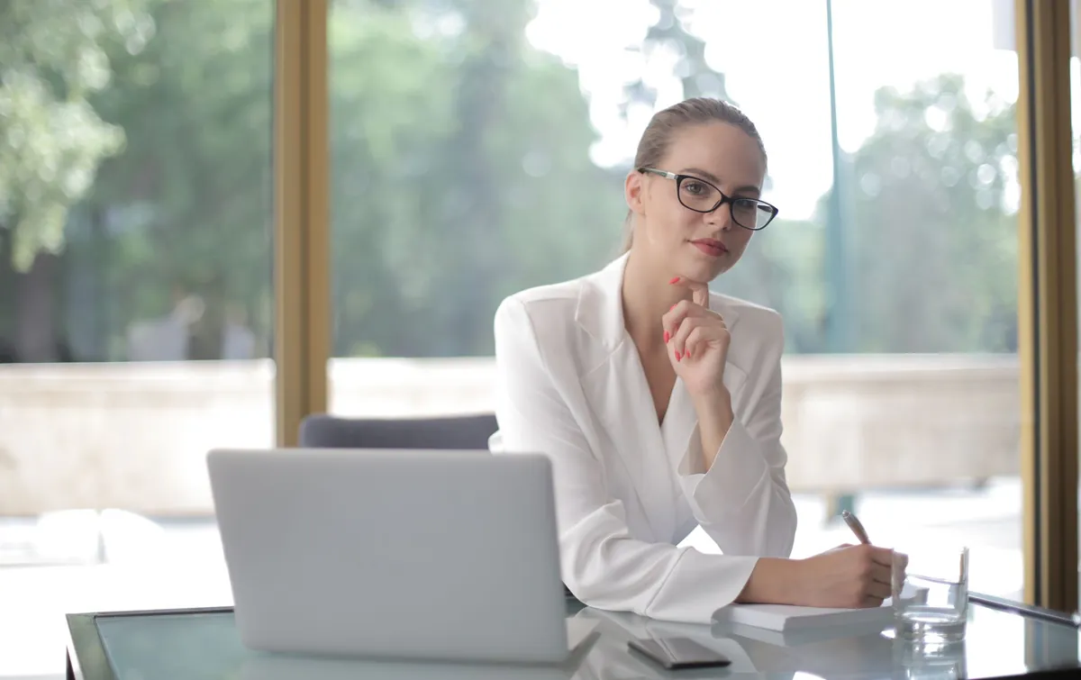 A woman in glasses sitting at a bright desk with a laptop, writing on a pad