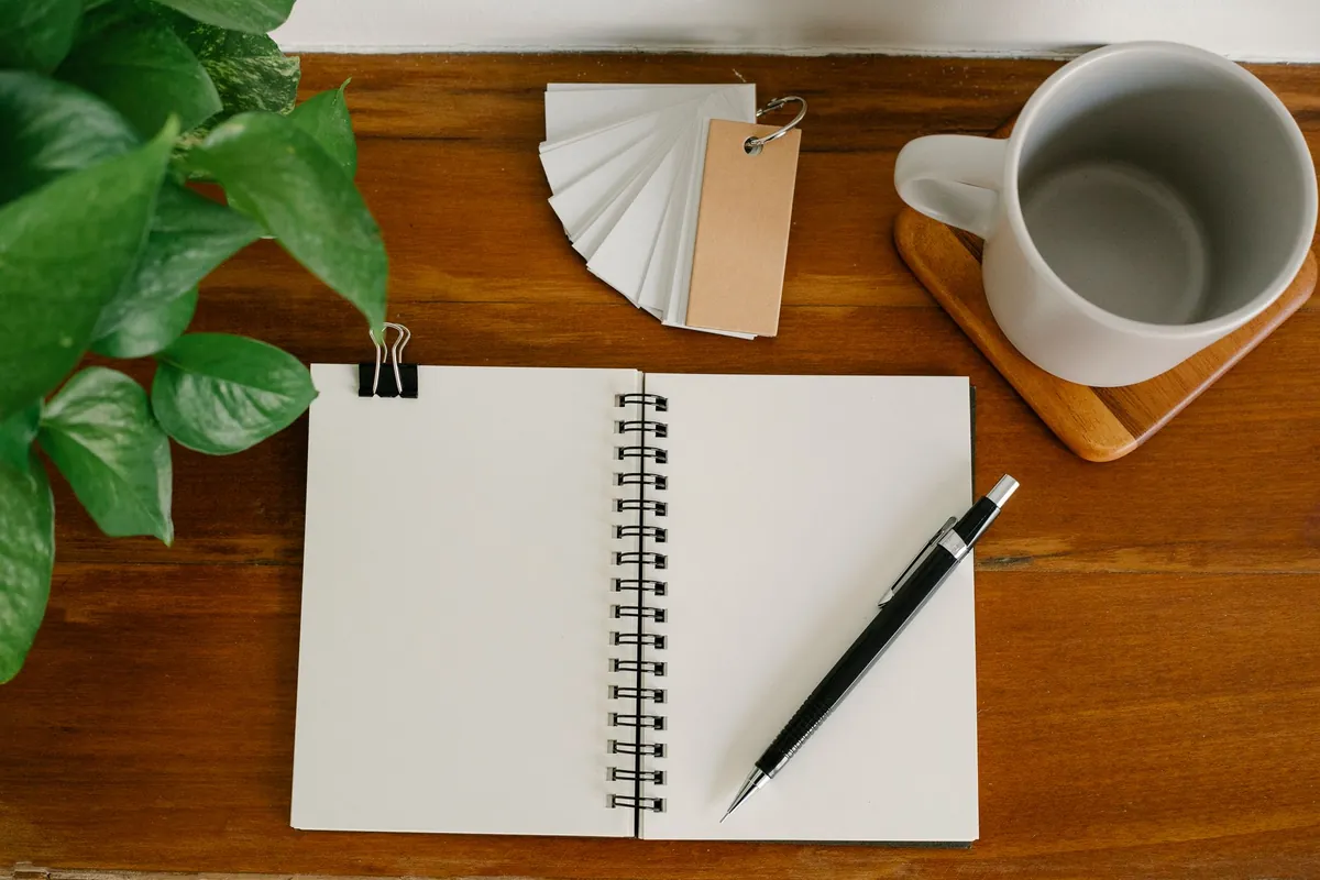 An open blank notebook with a pen, a mug and paper swatches on a wooden desk