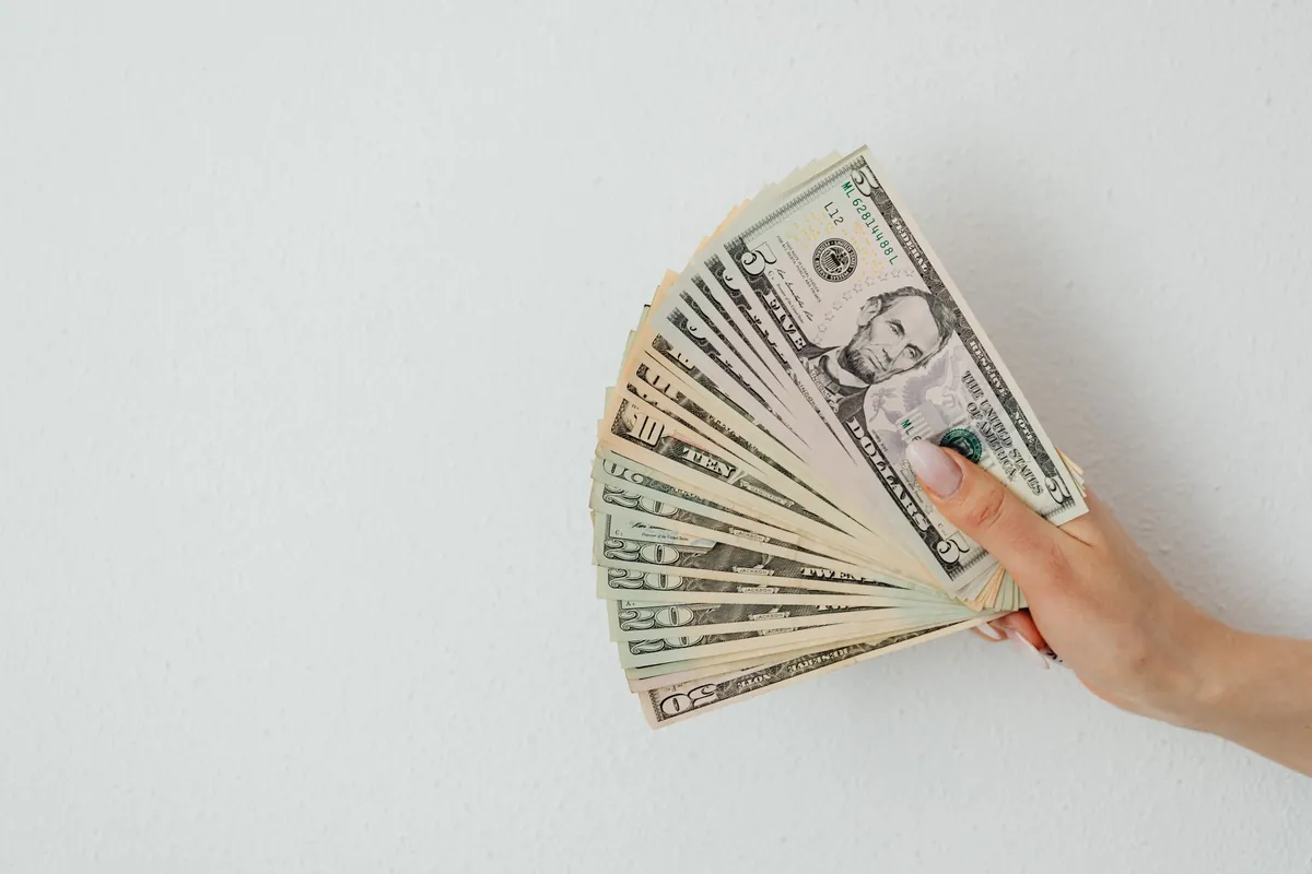 A hand holding a fan of dollar notes against a white wall