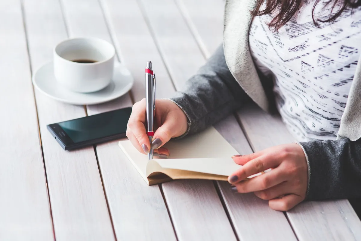 A woman writing in a journal beside a cup of coffee on a wooden desk