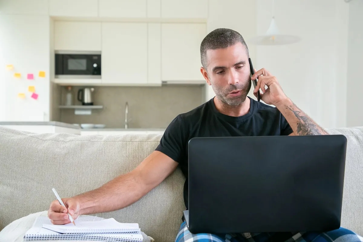 A man on the phone on a sofa, a laptop on his knees, writing notes