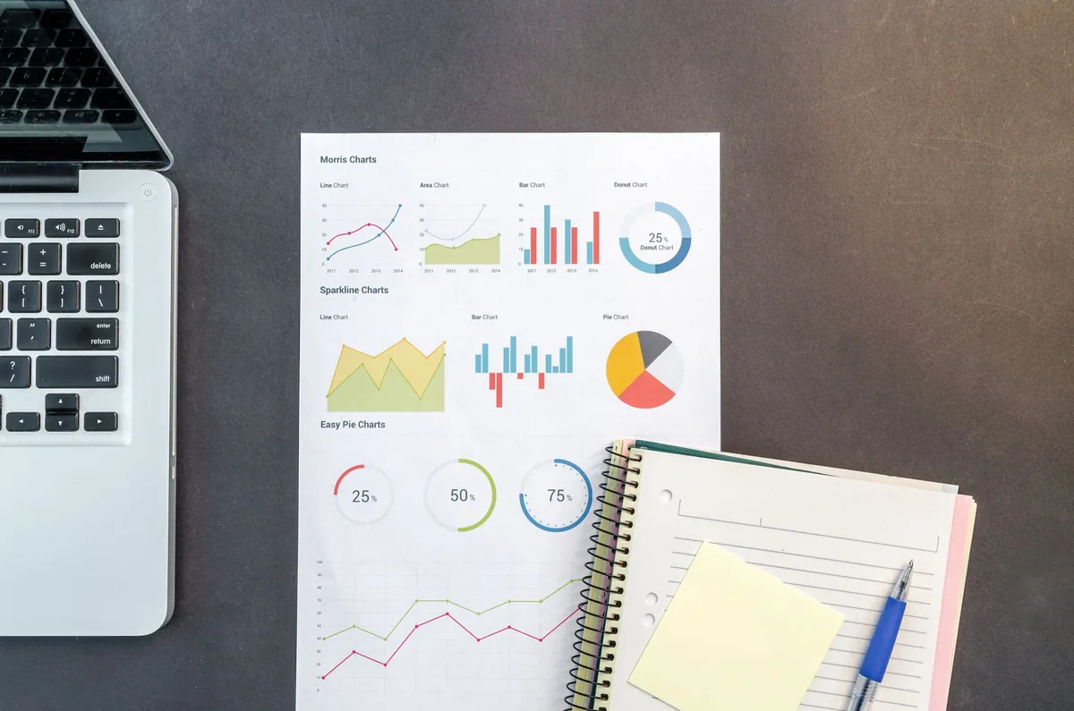 Printed charts and an open notebook on a dark desk beside a laptop