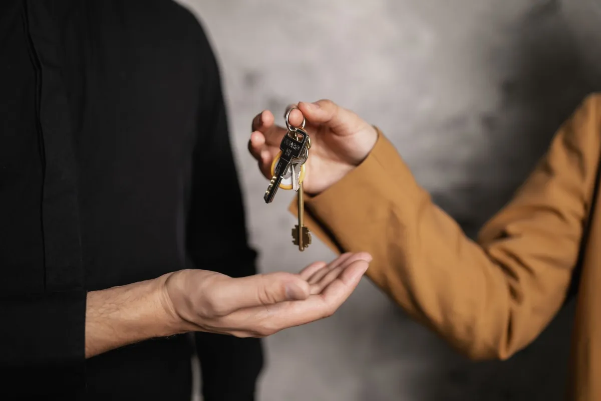 One hand holding a bunch of keys on a ring above another open upturned palm, against a plain grey concrete wall.