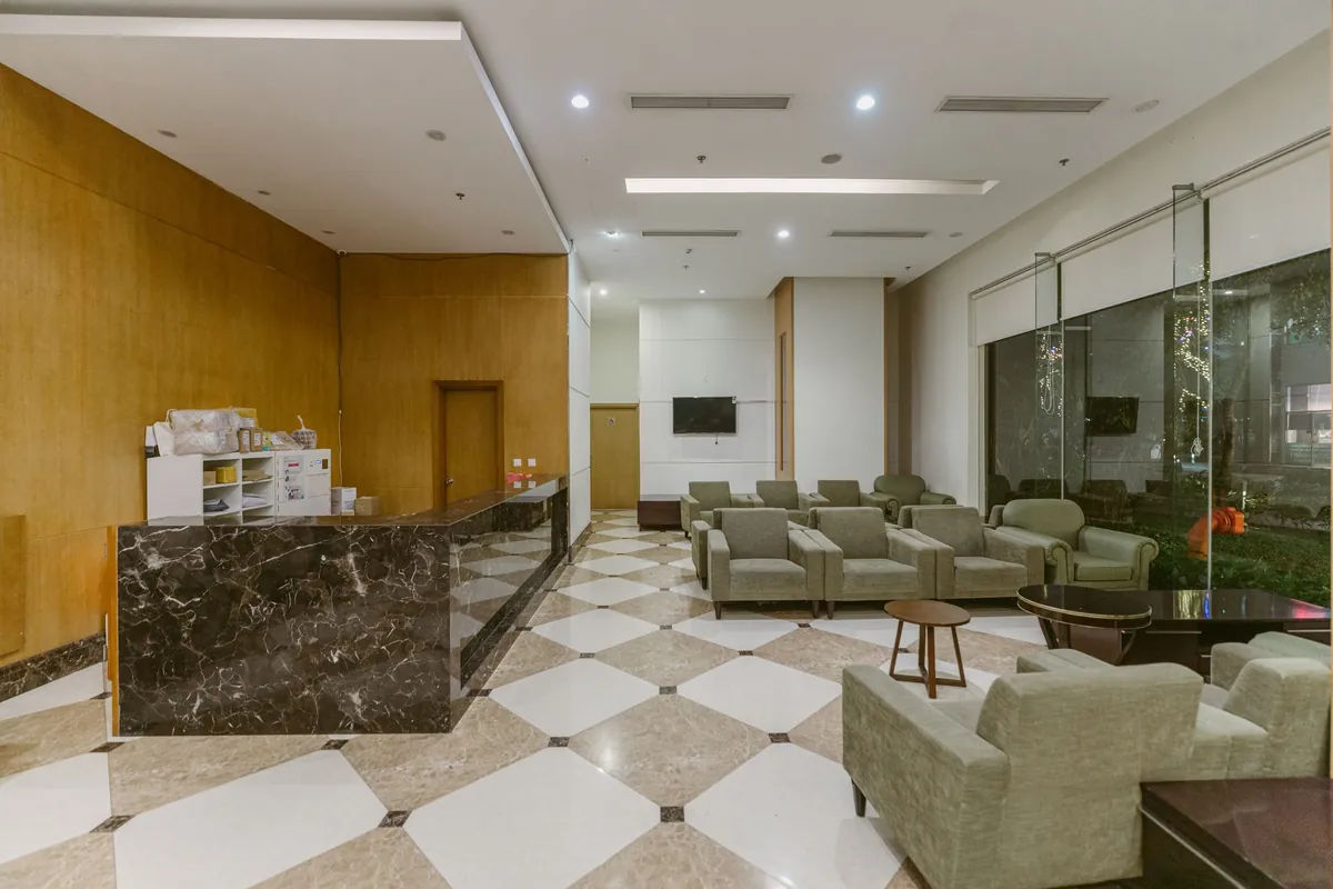 An empty hotel lobby with a dark marble front desk on the left, a chequerboard tiled floor and a row of grey armchairs by tall windows on the right.