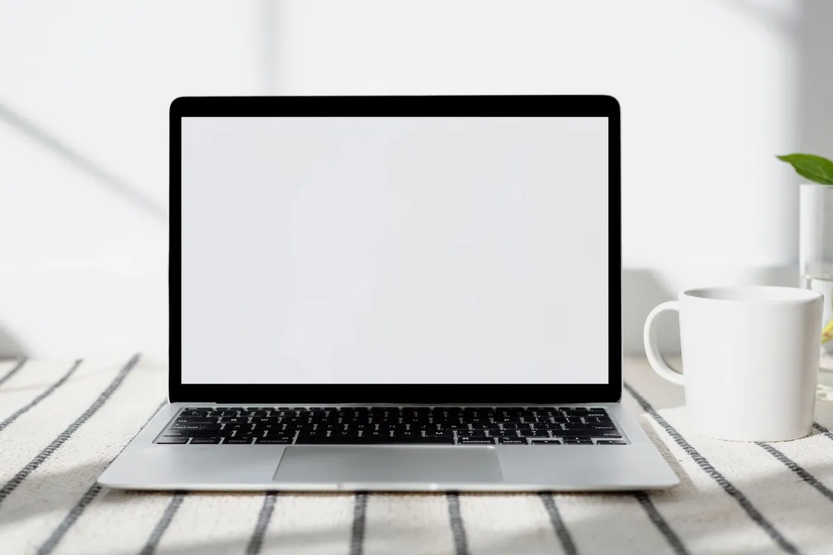 An open silver laptop with a blank white screen on a striped woven mat, a plain white mug and a glass vase beside it.