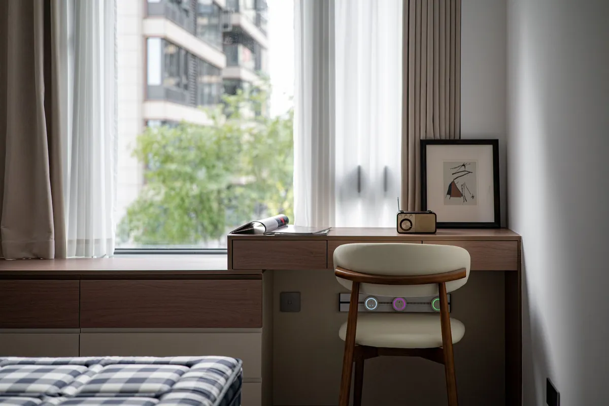 A wooden desk built under an apartment window with a chair, a small radio and a framed print, looking out to a neighbouring block and a tree.