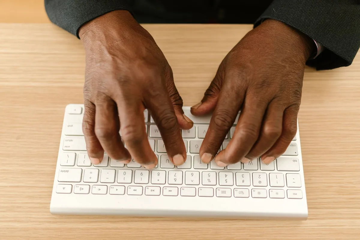 A pair of hands seen from behind and above, fingers resting on the keys of a slim white wireless keyboard on a light wooden desk.