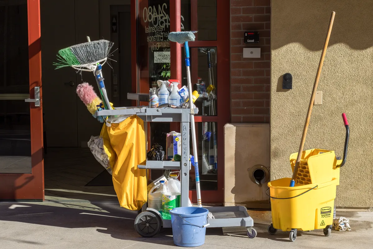 A cleaning cart with a mop bucket and supplies standing outside a building entrance in sunlight