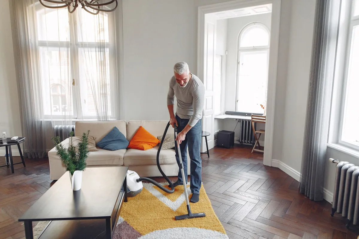 A person vacuuming the floor of a bright living room with tall windows