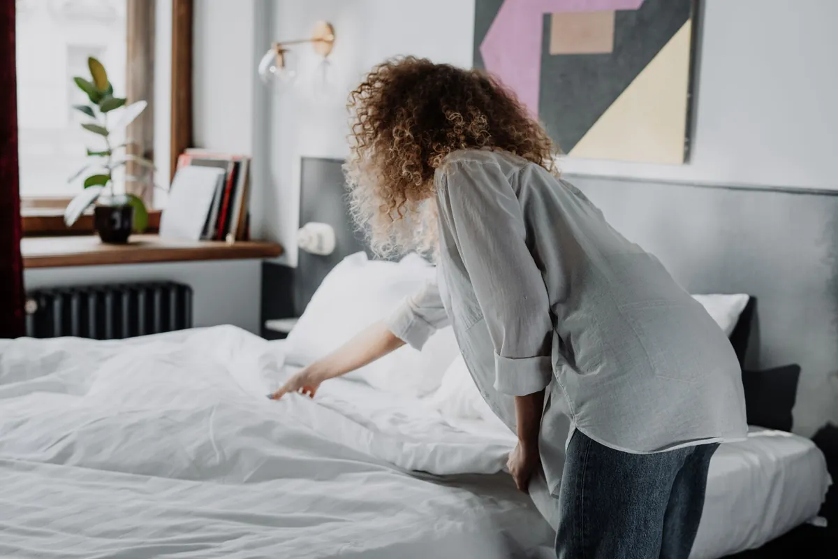 A woman making a bed with white linen in a modern bedroom with natural light