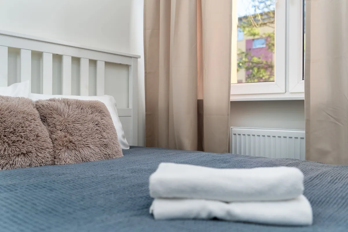 Two folded white towels laid on a grey waffle bedspread in a bright bedroom, with fluffy pillows, beige curtains and a radiator under the window.