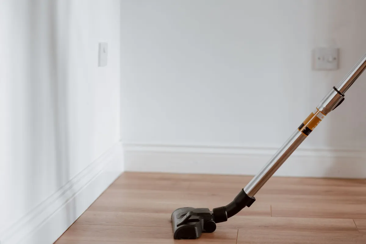 The head of an upright vacuum cleaner standing on a pale wood floor against a white skirting board in an empty, brightly lit room.