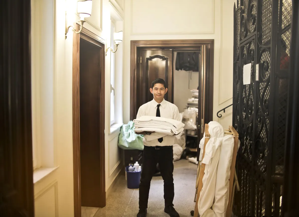 A man in a white shirt and tie carries a stack of folded white bed linen through a hallway, a wheeled linen rack and a laundry store behind him.