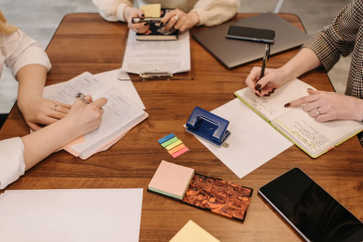 Hands of several people writing notes around a wooden table with phones and sticky notes