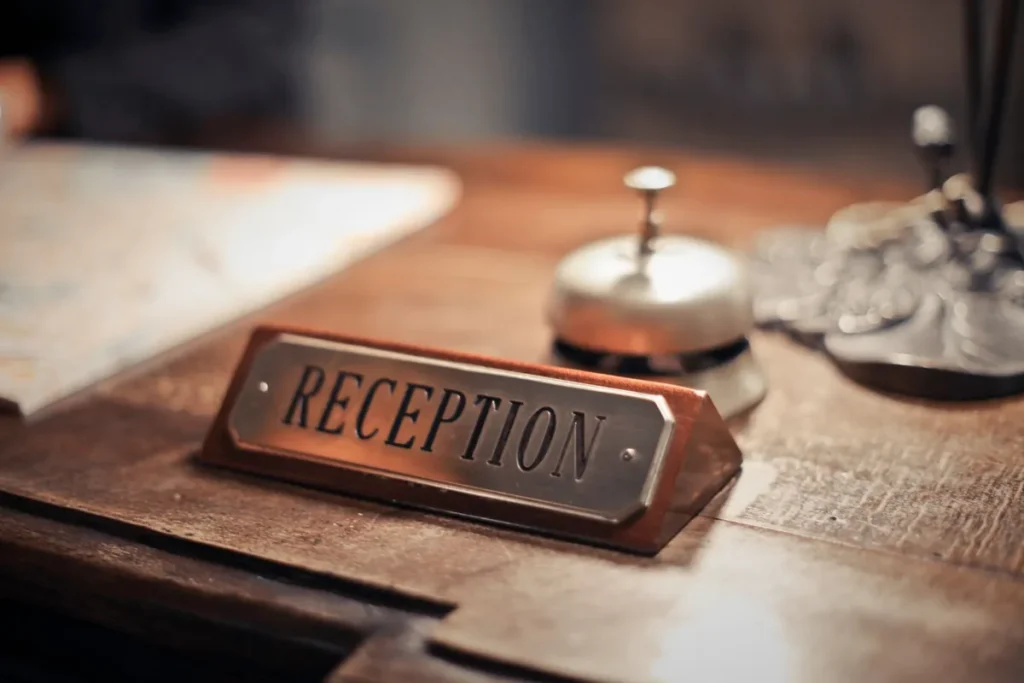 A brass RECEPTION desk plate in sharp focus on a worn wooden counter, with a silver service bell behind it and a folded map to the left.