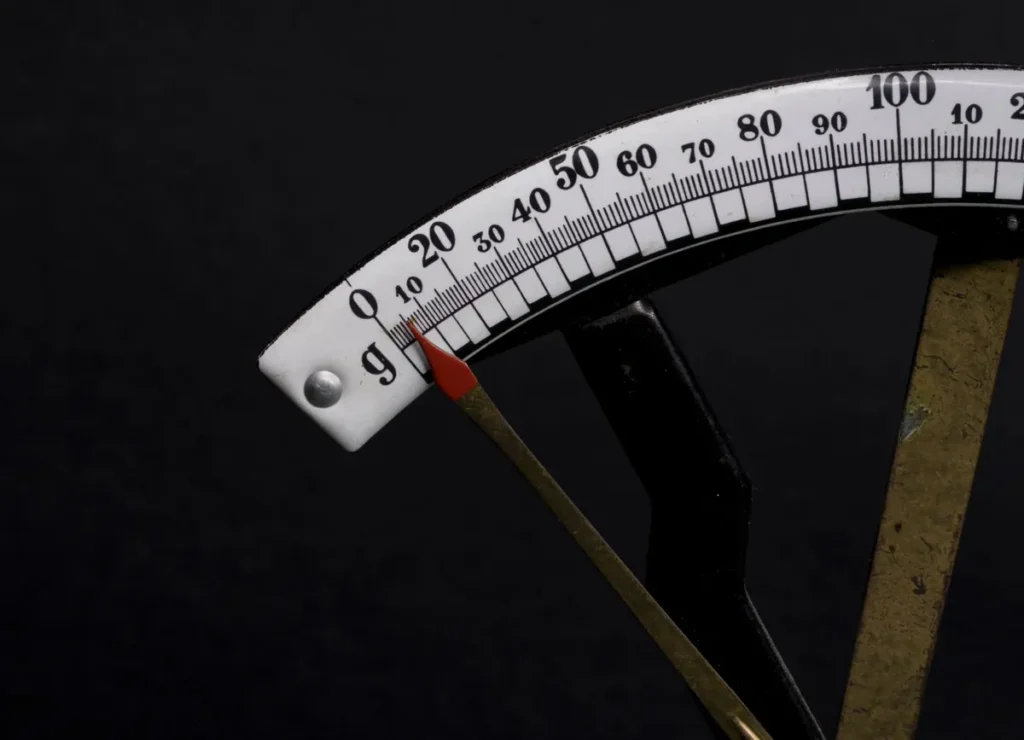 A macro of the curved white scale of a balance, its graduations running 0 to 100 in grams with a red pointer at zero, on a black background.