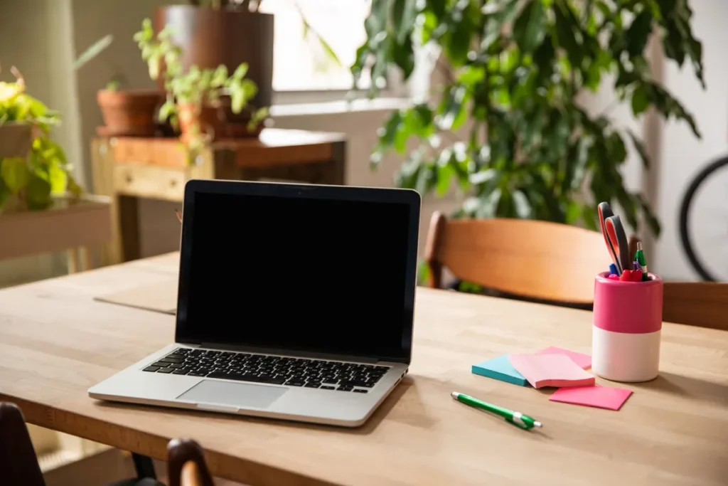 An open laptop with a blank dark screen on a wooden table beside sticky notes, a pen pot and house plants