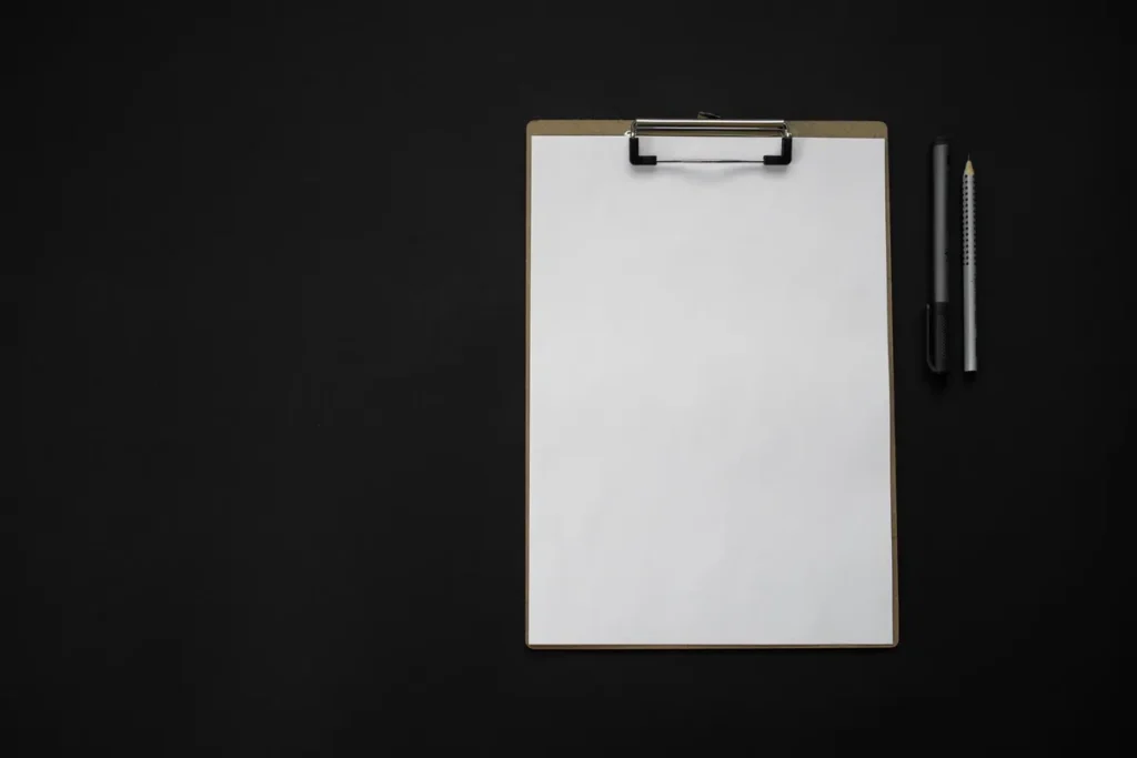 A blank clipboard holding a sheet of white paper, with a black pen and a pencil beside it, on a plain black background.