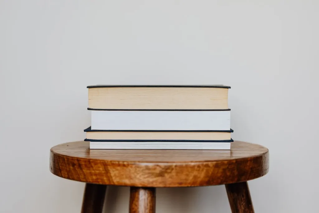 Three hardback books stacked neatly on a round wooden stool against a plain pale wall.