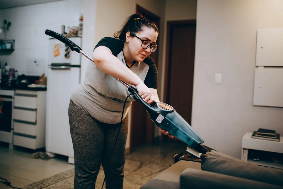 A woman vacuuming a sofa in a living room with the kitchen behind her