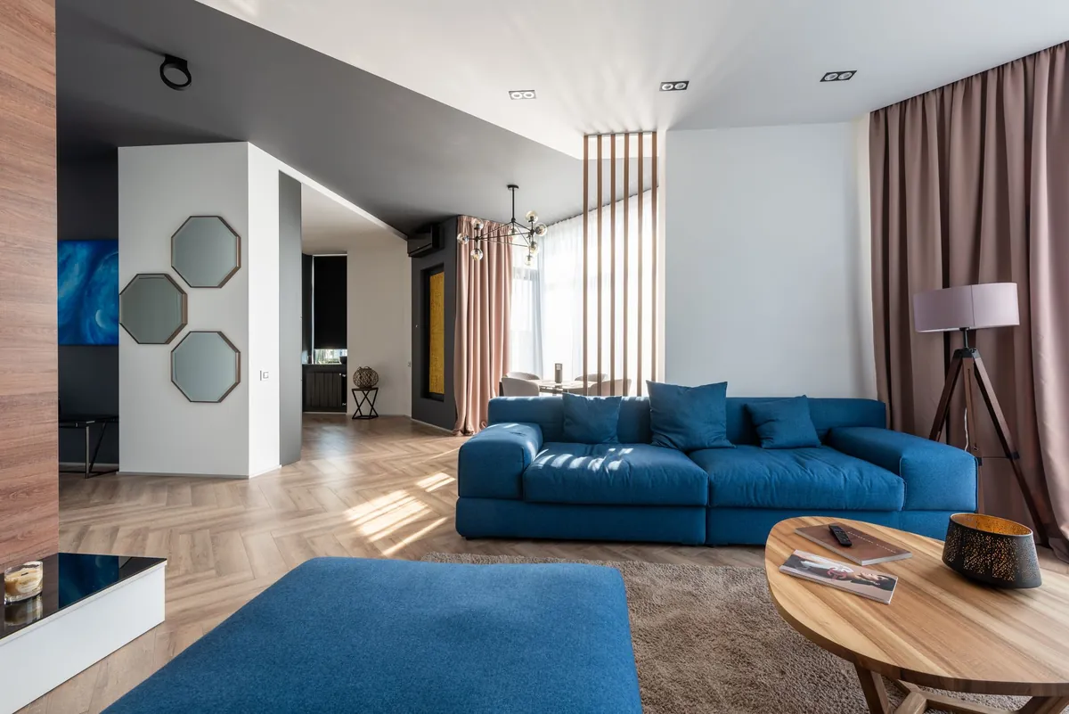 A tidy open-plan flat with a blue sofa and matching ottoman, herringbone wood floor, a tripod floor lamp and a wooden oval coffee table holding two books and a remote.