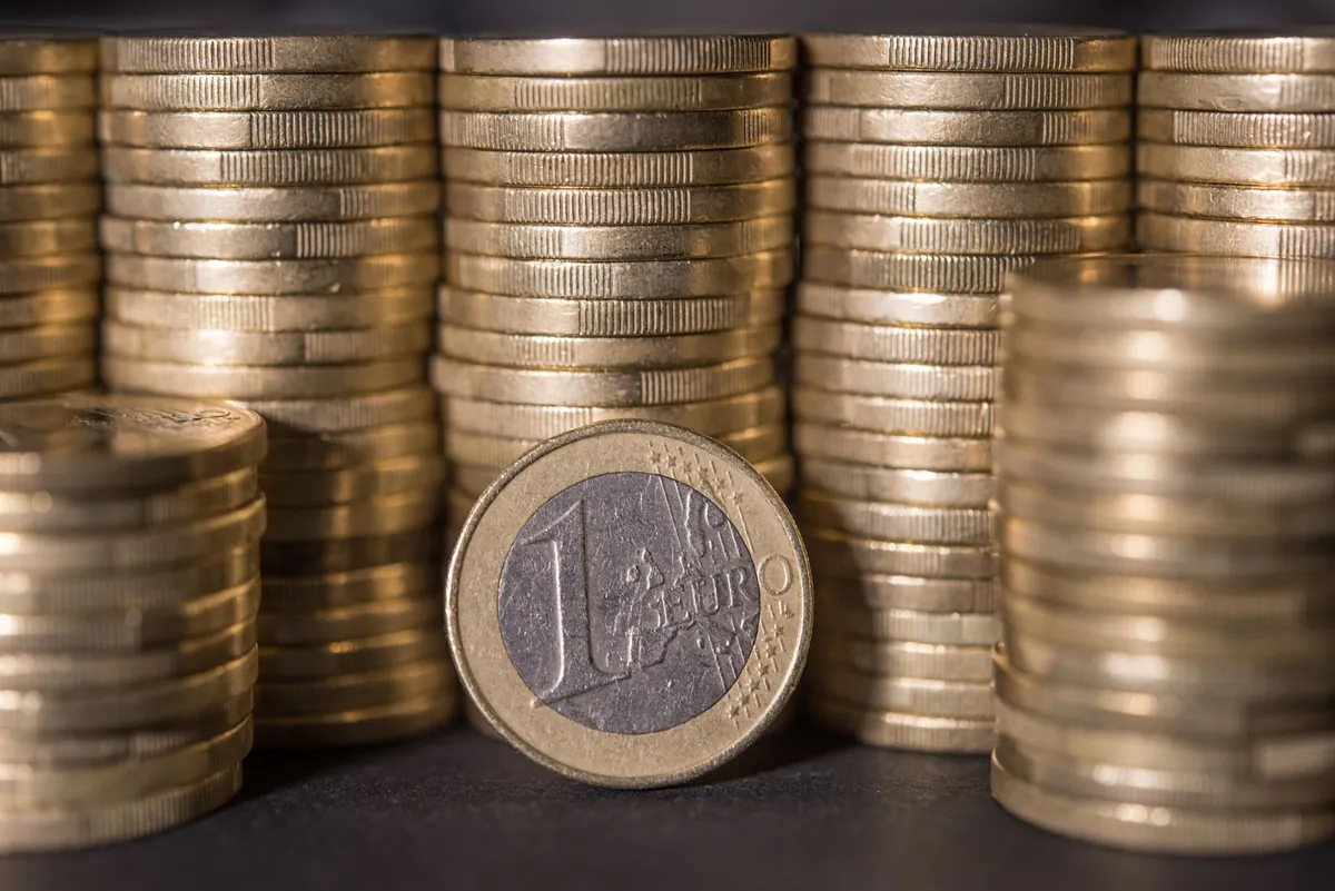 Tall stacks of gold-coloured euro coins filling the frame, with one 1 euro coin standing on edge showing its value face.