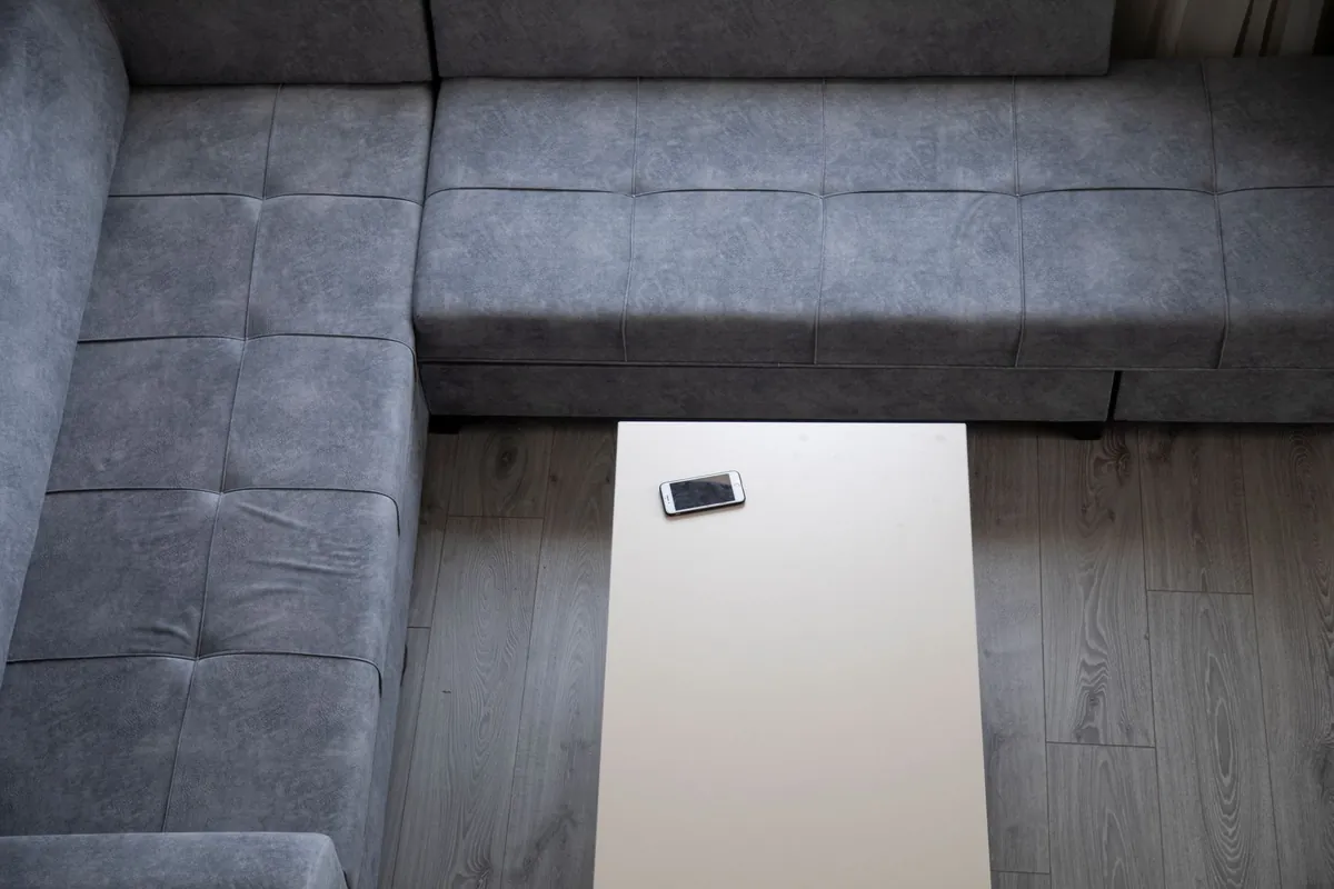 Looking down on a grey fabric corner sofa and a white rectangular coffee table with a single smartphone lying on it, on a grey wood floor.