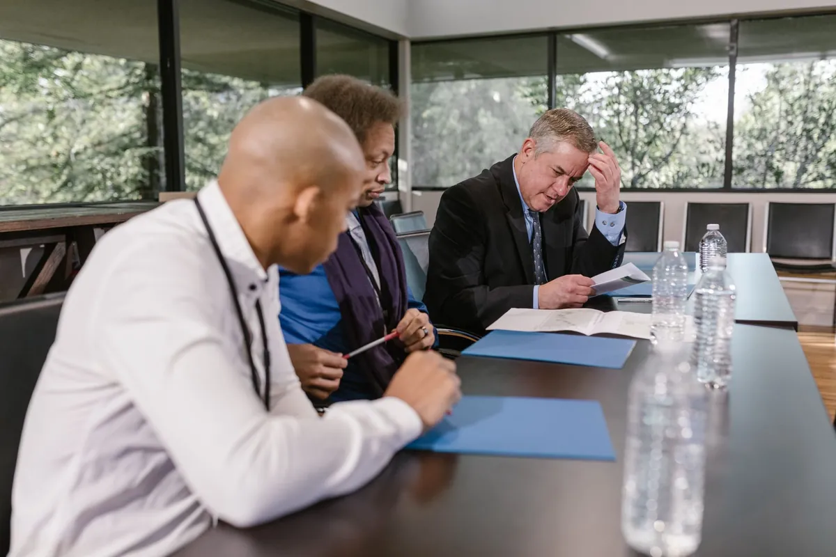 Three people along one side of a dark boardroom table, all looking down at papers and a folder, with clear water bottles in the foreground and trees through the window behind.