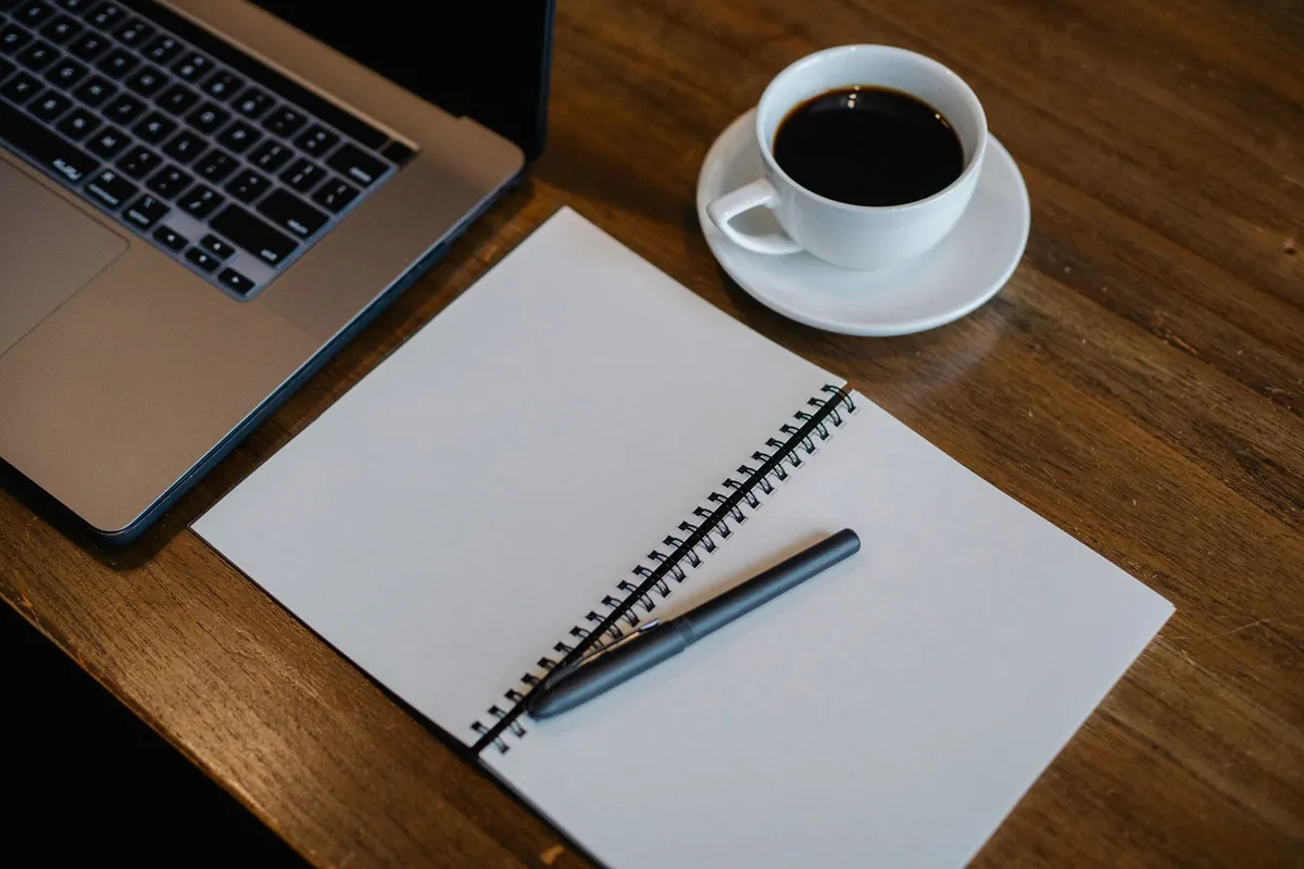An overhead view of a wooden desk with the corner of an open laptop, a blank spiral notepad with a pen laid across it, and a white cup of black coffee.