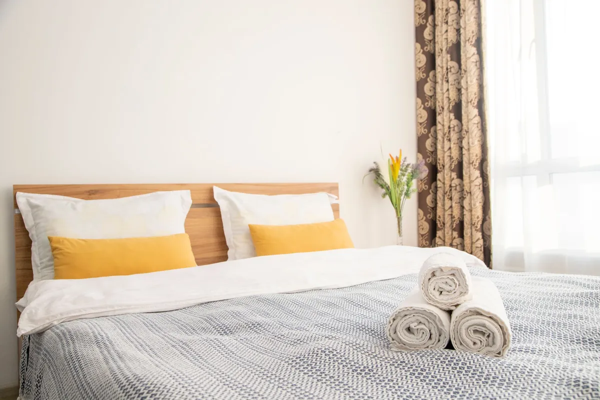 A made double bed with rolled towels, yellow cushions and a small vase of flowers