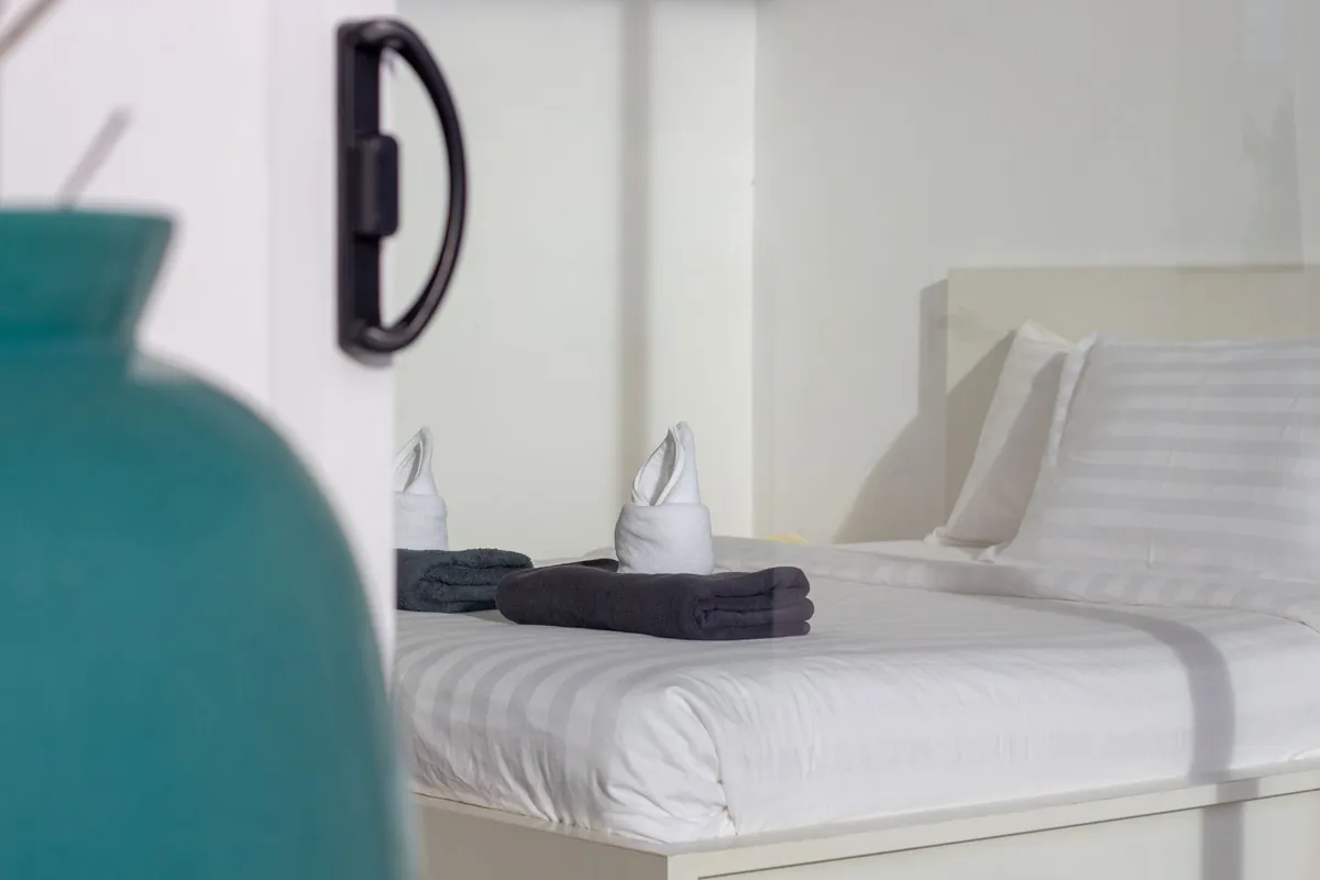 Folded towels set out on a made white bed next to a teal vase